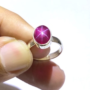Könnte beinhalten: Ein Silberring mit einem rosafarbenen Sternrubin-Edelstein. Der Rubin ist oval geformt und hat ein sternförmiges Muster.