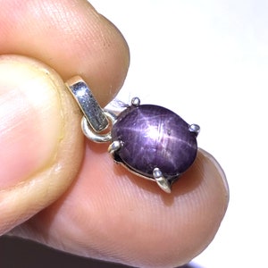 May include: A pendant with a dark purple star sapphire gemstone. The oval stone displays a six-ray star pattern, set in a silver metal setting with a loop for a chain.