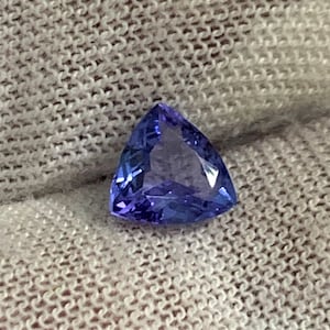 May include: A triangular cut, purple gemstone with a light blue hue. The gemstone is set on a white fabric background.