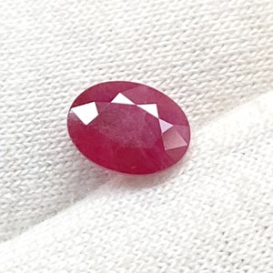 Könnte beinhalten: Ein lebhafter, ovaler Edelstein in Pink mit facettiertem Schliff. Der Edelstein liegt auf einem weißen Stoffhintergrund.
