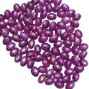 May include: A collection of oval-shaped, pink star ruby gemstones. The stones have a star-like pattern in the center and are arranged in a loose pile.