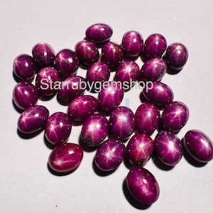 May include: A collection of oval-shaped, deep magenta star rubies, each featuring a distinct six-rayed star effect on its smooth, polished surface. The text "Starrubygemshop" is visible in the center of the arrangement of these loose gemstones.