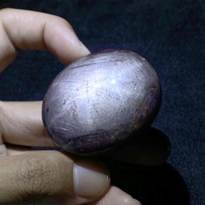 Könnte beinhalten: Ein polierter, oval geformter Sternrubin-Edelstein mit einem tiefen purpurroten Farbton. Die Oberfläche zeigt einen sichtbaren sechsstrahligen Sterneffekt, bekannt als Asterismus, der durch Lichtreflexion an internen Einschlüssen entsteht.