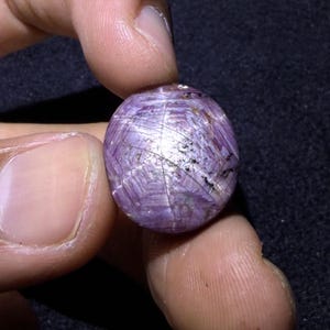 Könnte beinhalten: Ein runder, im Cabochon-Schliff gefertigter Sternrubin-Edelstein mit einem ausgeprägten sechsstrahligen Asterismus-Effekt auf seiner durchscheinenden, tiefvioletten und magentafarbenen Oberfläche, inklusive sichtbarer Einschlüsse.