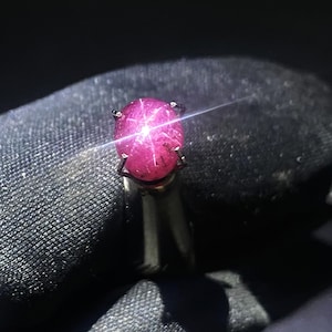 Könnte beinhalten: Ein Ring mit einem runden, im Cabochon-Schliff gearbeiteten Sternrubin in leuchtendem Pink-Rot. Der Edelstein zeigt bei direktem Lichteinfall einen deutlichen sechsstrahligen Sterneffekt und ist in einer einfachen Metallfassung mit Krappen gefasst.