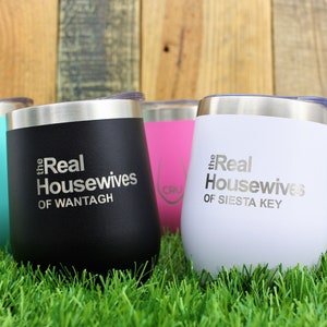 K&ouml;nnte beinhalten: F&uuml;nf Edelstahlbecher mit verschiedenen Farben f&uuml;r Deckel und K&ouml;rper. Die Becher tragen den Text "the Real Housewives" mit einem Ortsnamen darauf gedruckt. Die Becher stehen auf gr&uuml;nem Gras.