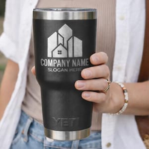 Puede incluir: Un vaso t&eacute;rmico negro con borde y base plateados. El vaso presenta un logotipo plateado con el texto "COMPANY NAME" y "SLOGAN HERE". La marca "YETI" est&aacute; impresa en la parte inferior.