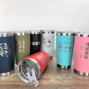 May include: Eight stainless steel tumblers with various colors and personalized engravings. The tumblers feature different designs and text, including "Best Dad Ever", "Hubby", "Elizabeth", "Vanessa", and "This Might Be Vodka".