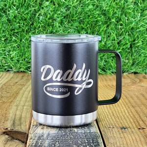Puede incluir: Taza de viaje negra con asa y tapa transparente. La taza tiene la palabra "Daddy" en escritura cursiva, con "SINCE 2021" debajo. La parte inferior de la taza es plateada.