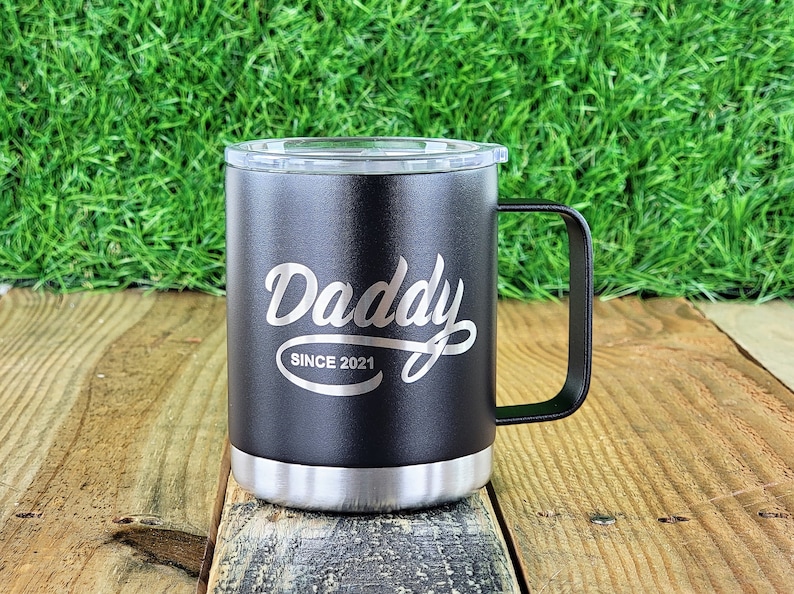 Puede incluir: Taza de viaje negra con asa y tapa transparente. La taza tiene la palabra "Daddy" en escritura cursiva, con "SINCE 2021" debajo. La parte inferior de la taza es plateada.