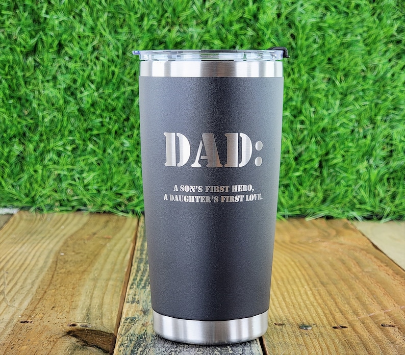 Puede incluir: Un vaso t&eacute;rmico negro con borde y tapa plateados. El vaso tiene la inscripci&oacute;n "DAD:" y el texto "A SON'S FIRST HERO, A DAUGHTER'S FIRST LOVE."