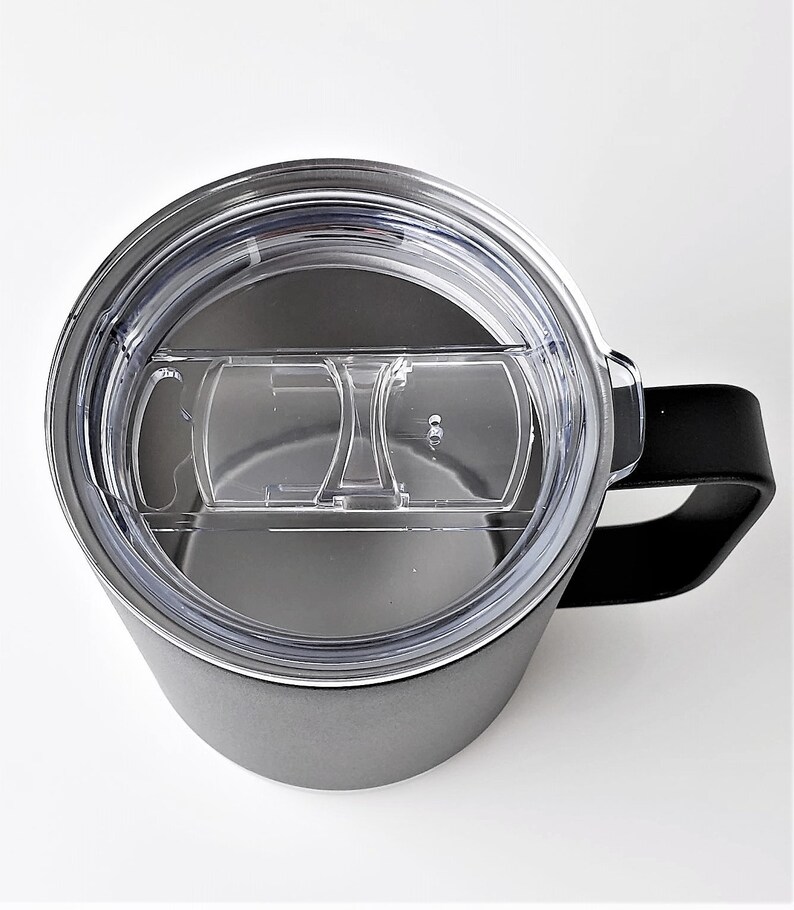 May include: A gray insulated travel mug with a black handle and a clear plastic lid. The lid has a sliding closure to open and close the drinking spout. The mug is designed to keep beverages hot or cold.