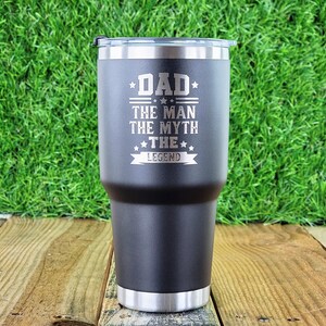 Pode incluir: Um copo t&eacute;rmico preto com aro e tampa prateados. O copo tem as palavras "DAD THE MAN THE MYTH THE LEGEND" gravadas na frente. O copo &eacute; cil&iacute;ndrico e parece ser feito de a&ccedil;o inoxid&aacute;vel.