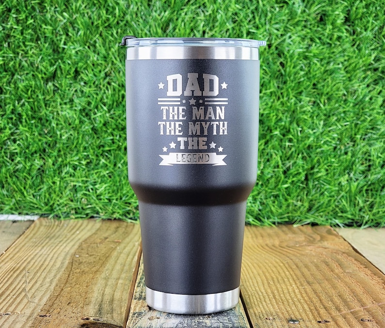 Pode incluir: Um copo t&eacute;rmico preto com aro e tampa prateados. O copo tem as palavras "DAD THE MAN THE MYTH THE LEGEND" gravadas na frente. O copo &eacute; cil&iacute;ndrico e parece ser feito de a&ccedil;o inoxid&aacute;vel.