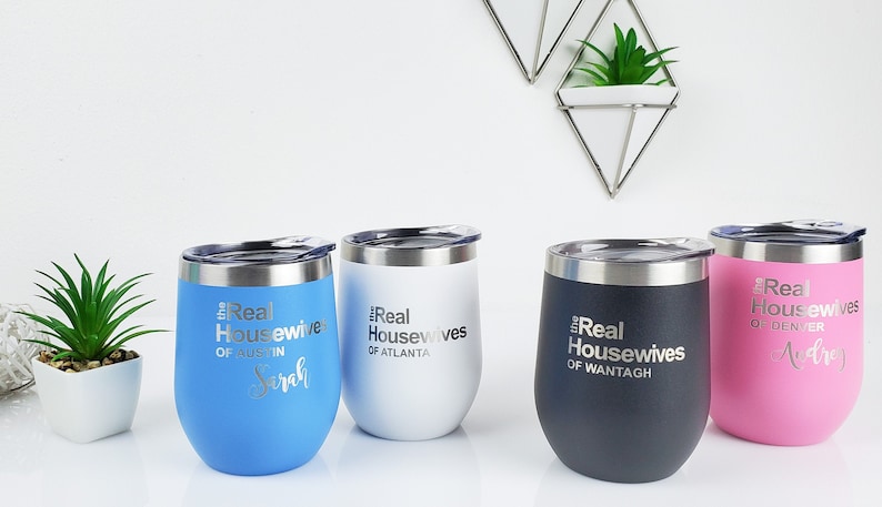 K&ouml;nnte beinhalten: Vier Edelstahl-Weingl&auml;ser mit Deckel. Jedes Glas hat eine andere Farbe und tr&auml;gt eine personalisierte Inschrift mit dem Text "The Real Housewives of" gefolgt von einem St&auml;dtenamen und einem Namen. Die Gl&auml;ser sind blau, wei&szlig;, schwarz und rosa.