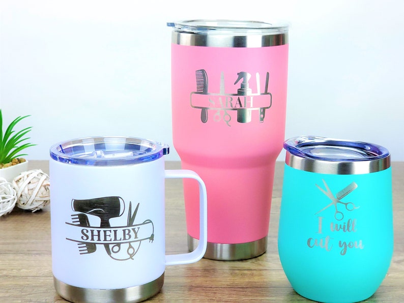 Puede incluir: Tres vasos de acero inoxidable con grabados personalizados para peluqueros. El vaso rosa dice "SARAH" con herramientas de peluquer&iacute;a. El vaso blanco dice "SHELBY" con un secador de pelo, tijeras y peines. El vaso turquesa dice "Te voy a cortar" con tijeras y una navaja.