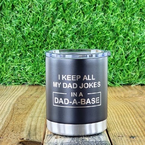 Puede incluir: Vaso negro con base plateada y tapa transparente. El vaso tiene el texto "I KEEP ALL MY DAD JOKES IN A DAD-A-BASE" en blanco. El vaso est&aacute; sobre una superficie de madera con un fondo de hierba verde.