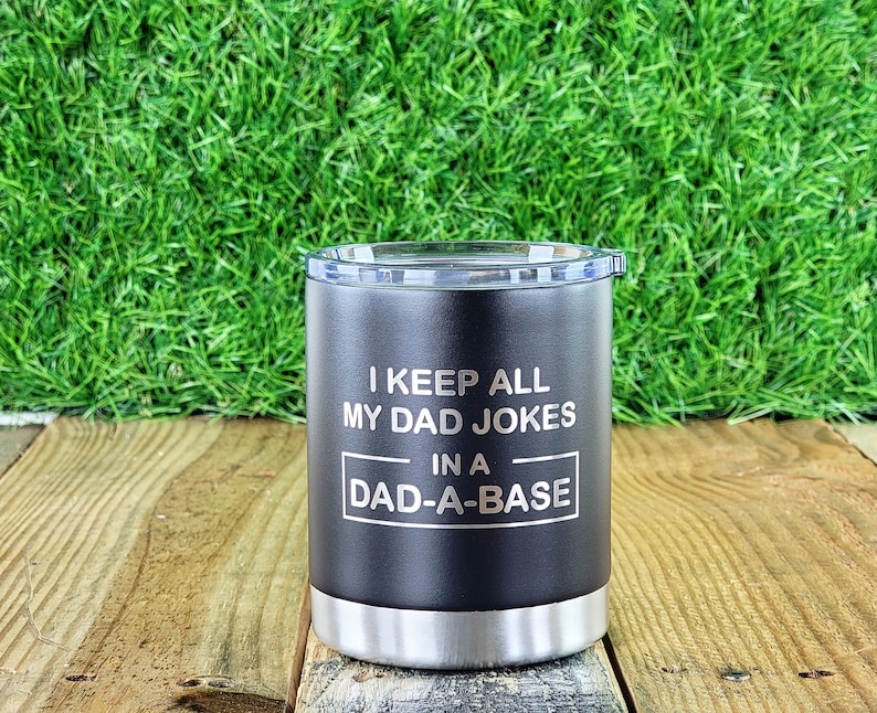 Puede incluir: Vaso negro con base plateada y tapa transparente. El vaso tiene el texto "I KEEP ALL MY DAD JOKES IN A DAD-A-BASE" en blanco. El vaso est&aacute; sobre una superficie de madera con un fondo de hierba verde.