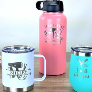 Puede incluir: Tres vasos de acero inoxidable con grabados personalizados. Una botella de agua rosa con el nombre "Charlotte" y herramientas de peluquer&iacute;a, una taza blanca con el nombre "Shelby" y un secador de pelo, y un vaso turquesa con el texto "Te voy a cortar" y unas tijeras.