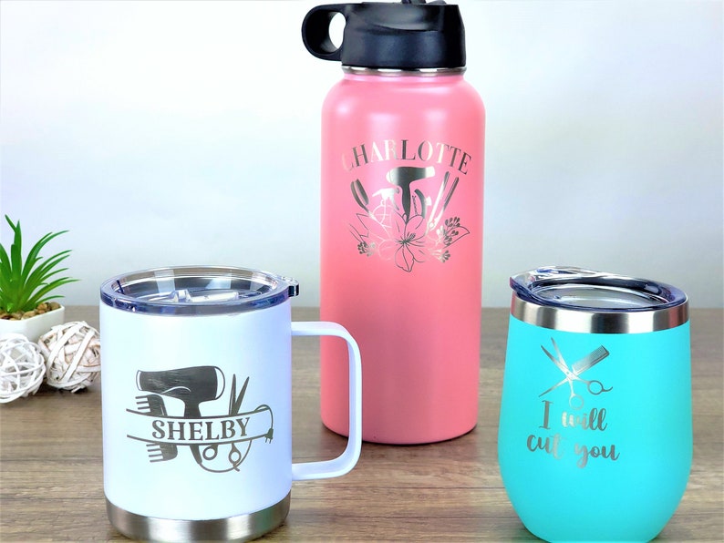 Puede incluir: Tres vasos de acero inoxidable con grabados personalizados. Una botella de agua rosa con el nombre "Charlotte" y herramientas de peluquer&iacute;a, una taza blanca con el nombre "Shelby" y un secador de pelo, y un vaso turquesa con el texto "Te voy a cortar" y unas tijeras.