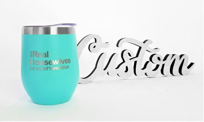 K&ouml;nnte beinhalten: Ein t&uuml;rkisfarbener Edelstahl-Weinglas mit dem Text "The Real Housewives of Chattanooga" eingraviert auf der Seite. Das Glas steht neben einem wei&szlig;en Holzschild mit der Aufschrift "Custom".