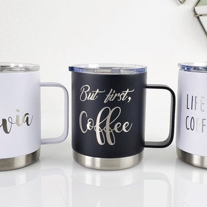 May include: Three travel mugs with stainless steel bases and handles. One white mug has the name "Olivia" in silver script. A black mug says "But first, Coffee" in silver script. A white mug says "Life happens, Coffee helps."