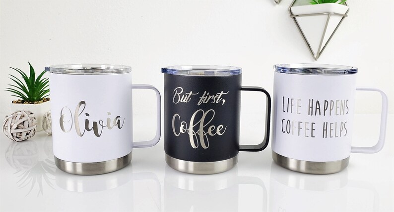May include: Three travel mugs with stainless steel bases and handles. One white mug has the name "Olivia" in silver script. A black mug says "But first, Coffee" in silver script. A white mug says "Life happens, Coffee helps."