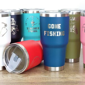 May include: A group of eight stainless steel tumblers with various designs and colors. The tumblers have a silver lid and a silver base. The designs include a stethoscope, a coffee cup, the words "Gone Fishing", a video game controller, and a floral design.