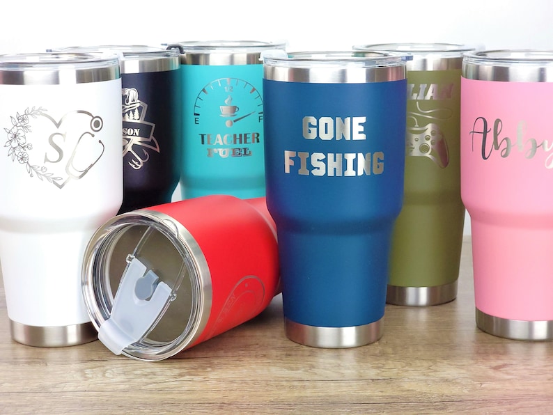 May include: A group of eight stainless steel tumblers with various designs and colors. The tumblers have a silver lid and a silver base. The designs include a stethoscope, a coffee cup, the words "Gone Fishing", a video game controller, and a floral design.