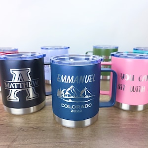 May include: A collection of insulated travel mugs in various colors, each with a handle and a lid. The mugs are engraved with names, initials, and phrases. One mug is navy blue with "EMMANUEL COLORADO 2022" and a mountain scene.
