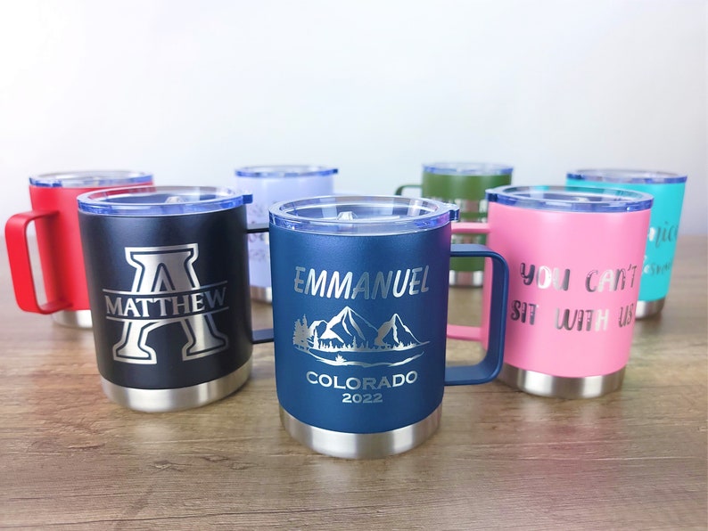 May include: A collection of insulated travel mugs in various colors, each with a handle and a lid. The mugs are engraved with names, initials, and phrases. One mug is navy blue with "EMMANUEL COLORADO 2022" and a mountain scene.