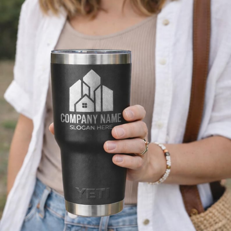 Puede incluir: Un vaso t&eacute;rmico negro con borde y base plateados. El vaso presenta un logotipo plateado con el texto "COMPANY NAME" y "SLOGAN HERE". La marca "YETI" est&aacute; impresa en la parte inferior.