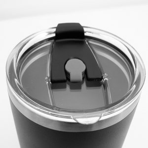 May include: Close-up of a black stainless steel travel mug with a clear lid and a black silicone slider.