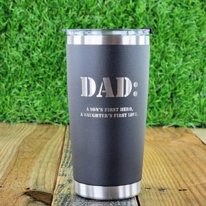 Pode incluir: Um copo t&eacute;rmico preto com borda e tampa prateadas. A palavra "DAD:" est&aacute; gravada no centro, com o texto "A SON'S FIRST HERO, A DAUGHTER'S FIRST LOVE" abaixo. O copo est&aacute; sobre uma superf&iacute;cie de madeira.