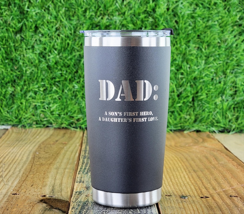 Pode incluir: Um copo t&eacute;rmico preto com borda e tampa prateadas. A palavra "DAD:" est&aacute; gravada no centro, com o texto "A SON'S FIRST HERO, A DAUGHTER'S FIRST LOVE" abaixo. O copo est&aacute; sobre uma superf&iacute;cie de madeira.