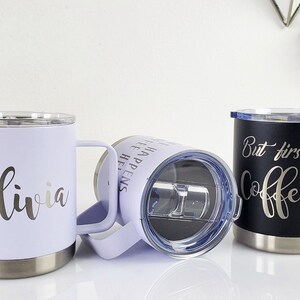 May include: Three insulated travel mugs with handles. One is lavender with "Olivia" in silver script, another is black with "But first, Coffee" in silver script, and the third is lavender with text. All have silver rims.