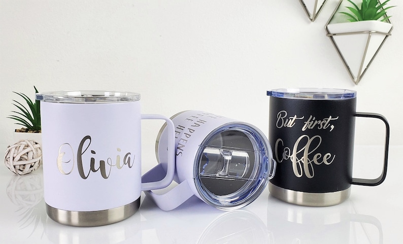 May include: Three insulated travel mugs with handles. One is lavender with "Olivia" in silver script, another is black with "But first, Coffee" in silver script, and the third is lavender with text. All have silver rims.