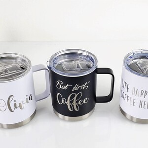 May include: Three insulated coffee mugs with handles and clear lids. One white mug has the name "Olivia" in script. A black mug has "But first, Coffee" in script. A white mug has "Life Happens Coffee Helps" in script.