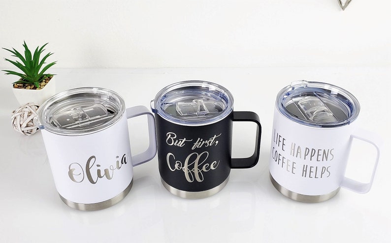 May include: Three insulated coffee mugs with handles and clear lids. One white mug has the name "Olivia" in script. A black mug has "But first, Coffee" in script. A white mug has "Life Happens Coffee Helps" in script.
