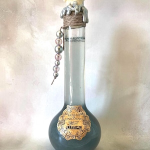 Op de afbeelding: Een glazen fles met een zilveren en gouden etiket waarop "Sangue di Unicorno Unicorn Blood ILLEGAL" staat. De fles is gevuld met een donkerblauwe vloeistof en heeft een zilveren en bruine dop met een kralenketting die eraan hangt.