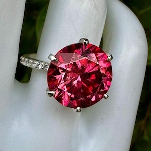 May include: A silver-toned ring featuring a large, round, faceted deep pink gemstone held by six prongs. The band is slender and decorated with a row of small, clear, sparkling stones along the side, creating a bright and reflective appearance.