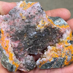 May include: A geode with a broken open cavity revealing a colourful interior. The interior is filled with small, white, pink, and yellow crystals. The exterior of the geode is a dark grey, green, and orange colour.