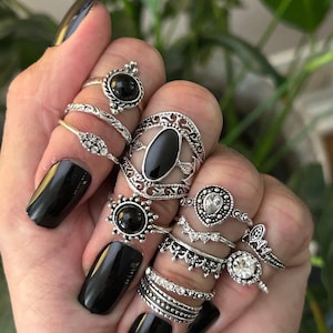 Puede incluir: Un conjunto de nueve anillos de plata con piedras negras y transparentes. Los anillos presentan diseños intrincados y están apilados en una mano con esmalte de uñas negro.