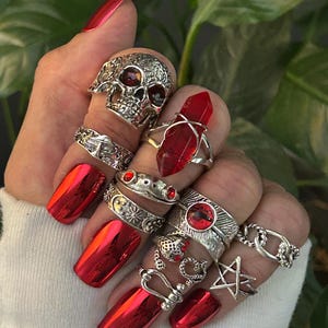 May include: A collection of silver rings with various designs, including a skull, a star, and a red crystal. The rings are worn on fingers with bright red, metallic nail polish. The overall aesthetic is gothic and edgy.