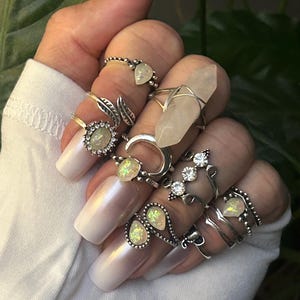May include: A hand wearing a variety of silver rings with opal and crystal accents. The rings feature different designs, including leaves, hearts, and geometric shapes.