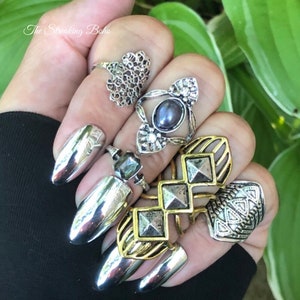 May include: A hand wearing several silver and gold rings. The rings feature various designs, including a heart, a flower, a geometric pattern, and a pyramid shape. The nails are painted a shiny silver colour.