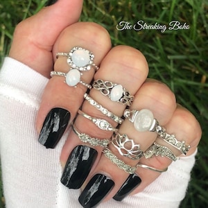 May include: A hand wearing multiple silver rings with white stones. The rings are stacked on each finger. The hand has black nail polish.