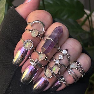 Może przedstawiać: Ręka z wieloma srebrnymi pierścionkami z opalem i kryształowymi akcentami. Pierścionki są ułożone na każdym palcu i zawierają pierścionek z fioletowym ametystem.