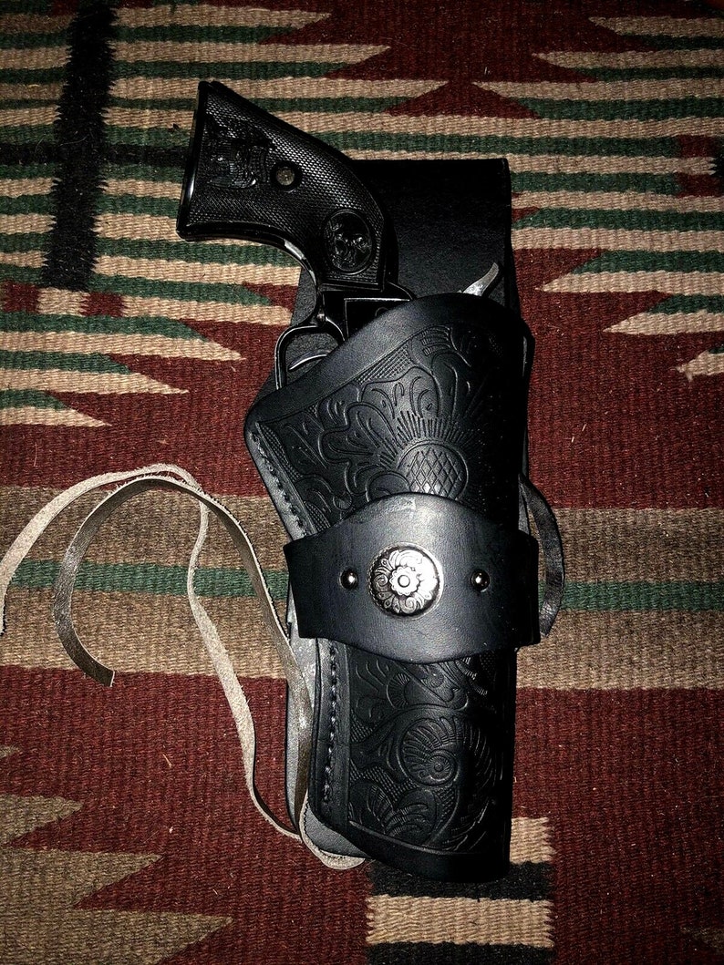 Peut inclure: Un &eacute;tui en cuir noir avec des motifs floraux complexes et un bouton argent&eacute;. L'&eacute;tui est con&ccedil;u pour contenir un revolver noir.