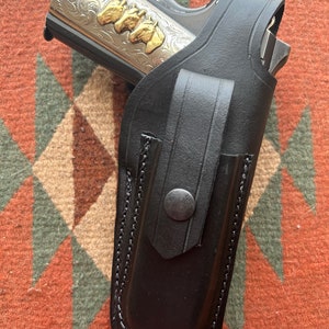 May include: A black leather holster with a stitched edge and a snap closure. A silver and gold handgun with an engraved design is inside the holster.